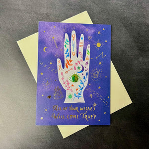 A blue card with an illustration of a hand with gold foil text that reads "All of your wishes will come true" against a cream color envelope. Sold at Coyote Supply Co a BIPOC and LGBTQIA+ owned zero waste witch store in Reno Nevada 