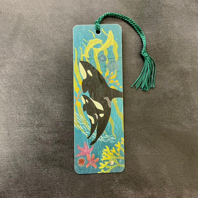 A wooden blue bookmark with a teal tassel featuring an illustartion of an orca and baby orca swimming among marine life. Sold at Coyote Supply Co a BIPOC and LGBTQIA+ owned zero waste witch store in Reno Nevada