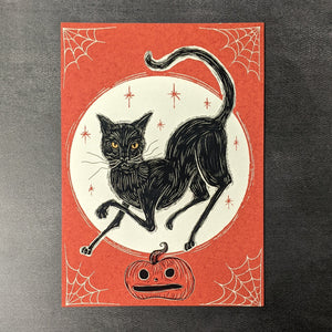 A print of an illustration of a black cat in a white circle against an orange background, below the cat s a jack o lantern and spiderwebs in each corner of the image by Bat In Your Belfry. Sold at Coyote Supply Co a BIPOC and LGBTQIA+ owned zero waste witch store in Reno Nevada 