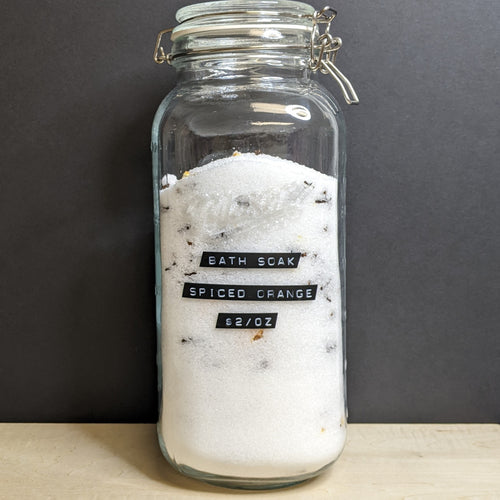 A tall glass jar full of white powder bath soak and spices with a black label that reads Bath Soak Spiced Orange.  Sold at Coyote Supply Co a BIPOC and LGBTQIA+ owned zero waste witch store in Reno Nevada 